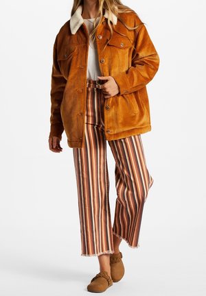 Kvinde iført en sennepsgul fløjlsjakke med hvid krave, stribede bukser med vide ben og brune mokkasinsko mod en hvid baggrund.