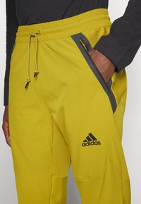 Gelbe Sporthose mit elastischem Bund, Kordelzug, seitlichen Reißverschlusstaschen und schwarzem Logodetail. Strukturierte Akzente an den Seiten.