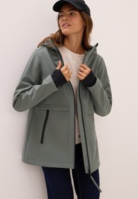 Veste softshell de couleur vert muted avec fermeture éclair, capuche et accents de poignet noirs. Elle dispose de poches latérales et d'une coupe droite.
