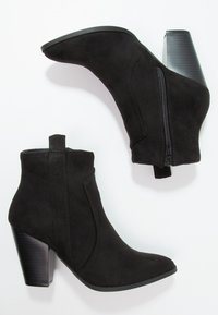 Czarne zamszowe botki za kostkę z bocznym zamkiem błyskawicznym i grubymi obcasami typu słupek, przedstawione na białym tle.