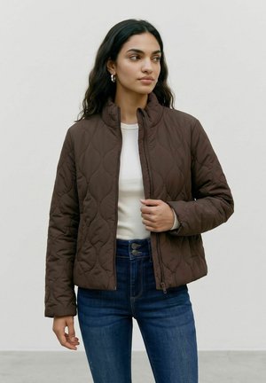 Femme aux longs cheveux foncés portant des boucles d’oreilles créoles argentées, une veste matelassée marron sur une chemise blanche et un jean bleu, debout devant un fond uni.