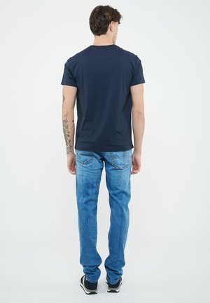 Man met armtattoo draagt een marineblauw T-shirt met korte mouwen, blauwe spijkerbroek en zwarte sneakers, staand met het gezicht naar een witte achtergrond.