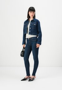 Woman wearing dark blue cropped denim jacket and skinny jeans, light gray top, black pointed heels, holding small black handbag.