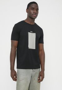 Čierne tričko s krátkym rukávom, na ktorom je umiestnená obdĺžniková grafika v sivej farbe s vertikálnymi pruhmi a nápisom "BOSS" hore. Materiál bavlna.