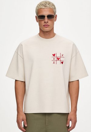 Blonder Mann mit schwarzen rechteckigen Sonnenbrillen und einem beigen T-Shirt mit rotem Tic-Tac-Toe-Design aus Herzen und X auf der Brust.