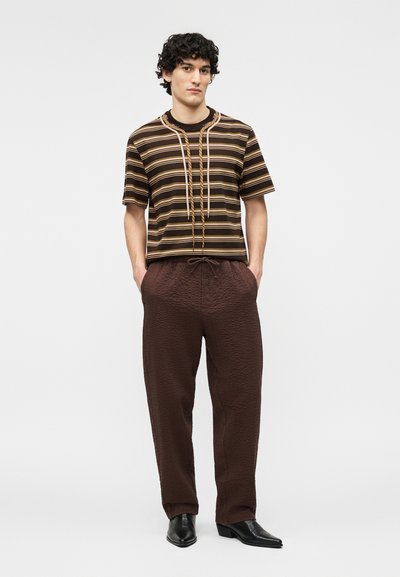 Jeune homme portant une chemise à manches courtes marron rayée et un pantalon marron foncé texturé, debout les mains dans les poches et chaussé de chaussures noires.