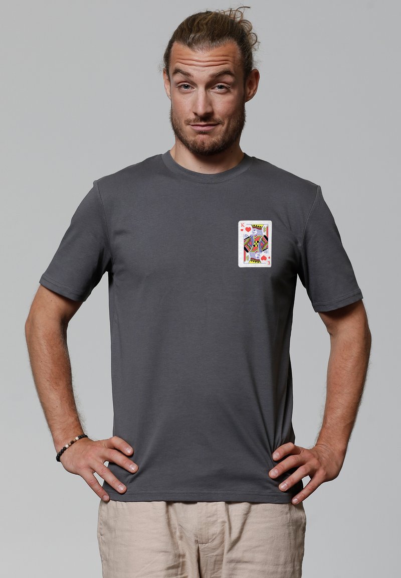 Jeune homme aux cheveux attachés, debout les mains sur les hanches, portant un t-shirt gris foncé avec un motif de carte à jouer Roi de Cœur.