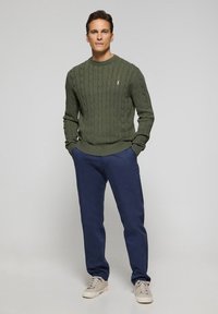 Olivgrön kabelstickad tröja, marinblå byxor och beige sneakers. Tröjan har en strukturerad design, klassisk rund halsringning och ribbade ärmslut.