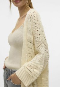 Cardigan lavorato a maglia color crema, caratterizzato da un design a maglia aperta con motivo testurizzato e polsini a coste. Indossato sopra una canottiera beige aderente.
