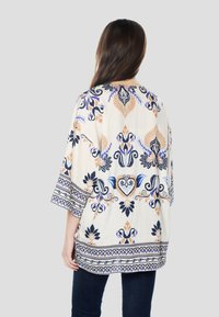 Izšūta kremkrāsas tunika ar zilu, tumši zilu un oranžu ziedu rakstiem; ar 3/4 garuma piedurknēm un izsmalcinātu apakšmalas dizainu.