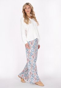 Vrouw in een lange mouwen witte blouse en wijde broek met blauw, oranje en witte geometrische patronen, die beige instapschoenen draagt en zijwaarts kijkt.