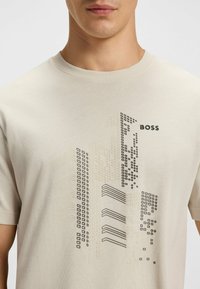 Beige puuvillane t-särk, millel on musta ja halli värvi graafika, mis kujutab hooneid, ning sõna "BOSS", mis on paigutatud ülemisse paremasse nurka.