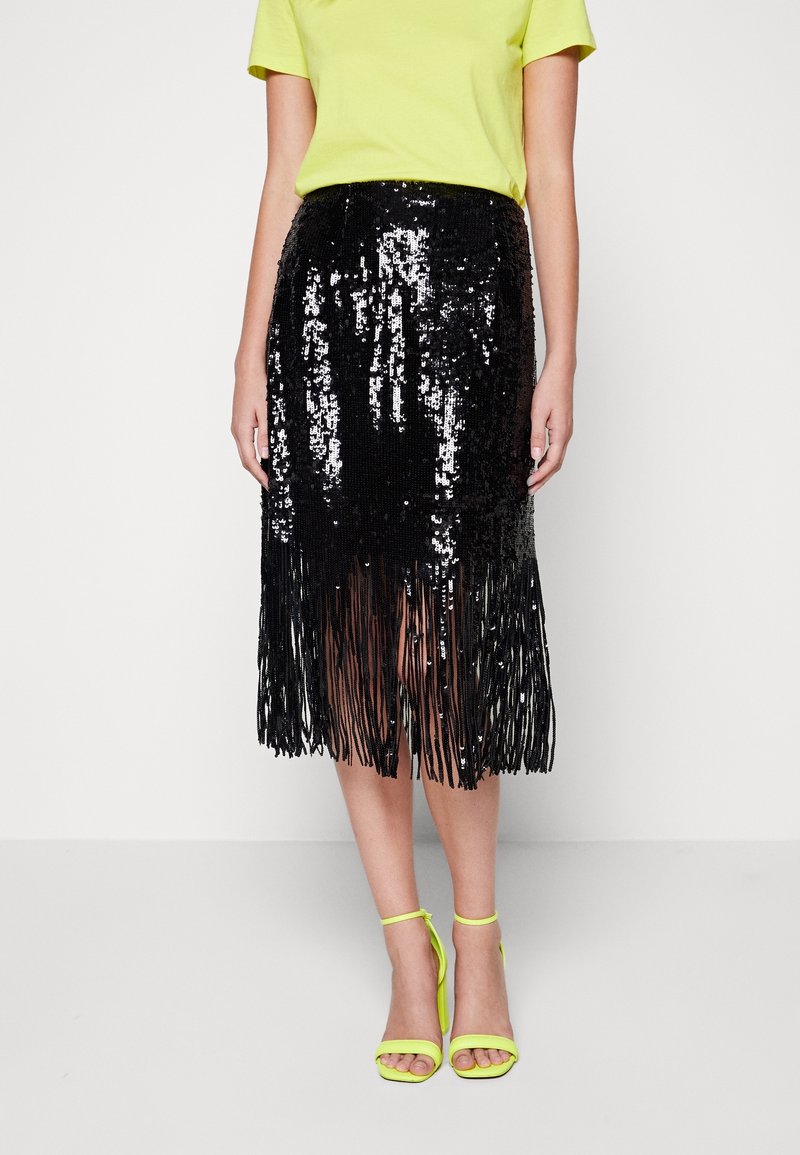 Femme portant une jupe à franges en sequins noirs, un haut à manches courtes vert citron et des sandales à talons hauts assorties vert citron, debout devant un fond blanc.