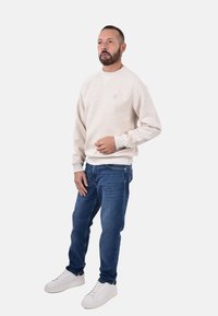 Felpa beige con collo rotondo e polsini a coste, abbinata a jeans blu e sneaker bianche. Design semplice, texture morbida.