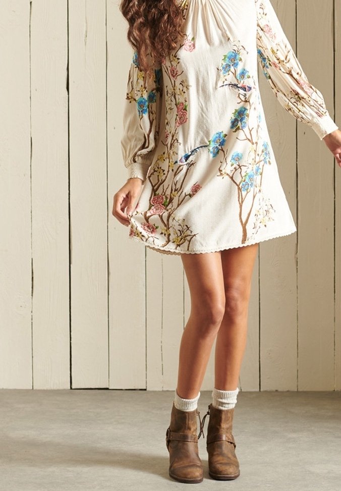 Femme portant une robe fleurie crème à manches longues avec des oiseaux, associée à des chaussettes blanches et des bottines marron, debout contre un mur en bois.