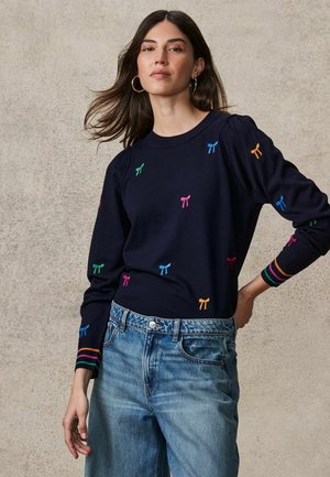 Navy-Pullover mit buntem Bogenstickerei, gerippten Bündchen mit Streifen. Kombiniert mit hochgeschnittenen hellblauen Jeans.