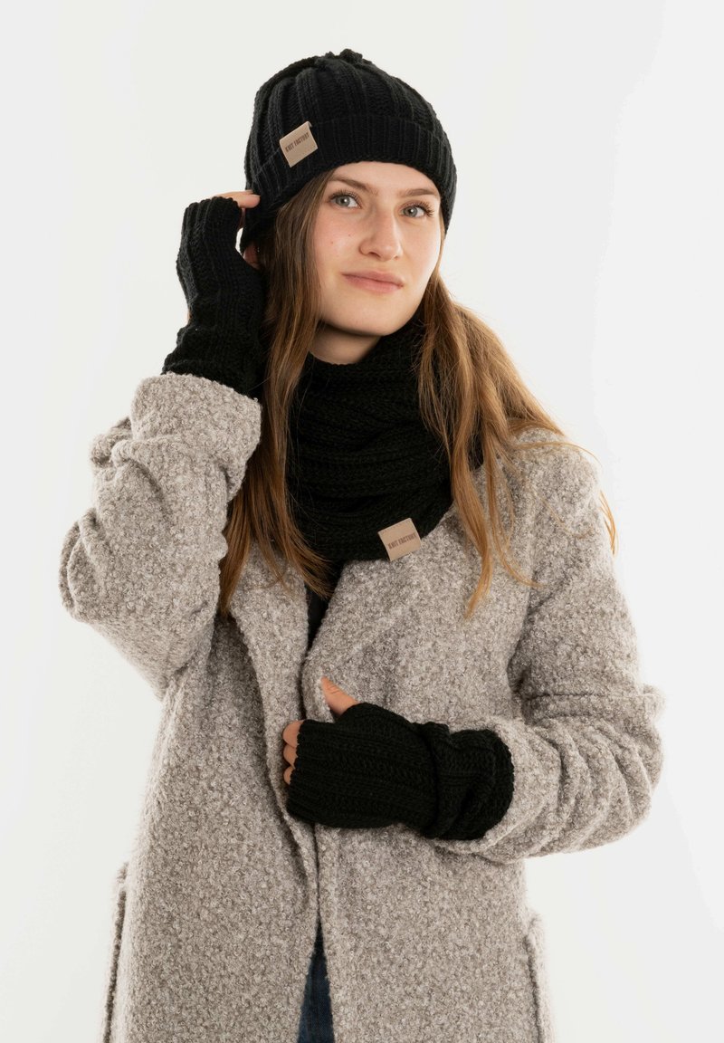 Zwarte gebreide muts, sjaal en handschoenen zonder vingers. Zachte textuur, geribbeld ontwerp, met een leren label op de sjaal. Gedragen bij een grijze jas.
