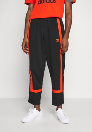 Hombre vistiendo pantalones deportivos Adidas negros con franjas laterales naranjas y zapatillas blancas, de pie contra un fondo claro y liso.