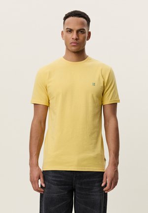 Jeune homme debout face à l'objectif, portant un t-shirt jaune uni à manches courtes et un jean foncé sur un fond clair.
