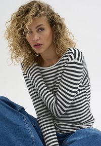 Striped long-sleeve shirt in white and navy, made from soft fabric. The model is wearing blue denim jeans, seated against a neutral background.