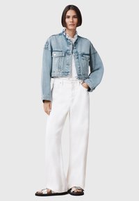 Chaqueta de mezclilla azul clara con diseño recortado, cuello deshilachado, dos bolsillos en el pecho; combinada con pantalones anchos blancos y sandalias negras.