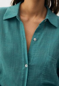 Teal button-up shirt with a collar, pocket detail, and pearlescent buttons; features a textured, slightly crinkled fabric.