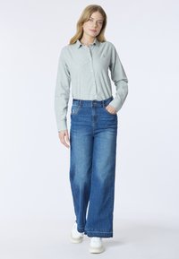 Camicia grigia chiaro, con bottoni e maniche lunghe; abbinata a jeans blu a vita alta e a gamba larga; scarpe da ginnastica bianche, look casual.