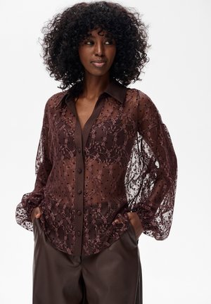 Durchsichtige burgunderfarbene Bluse mit Spitzen- und Punktmustern, langen Puffärmel und einem dunklen Kragen, kombiniert mit braunen Lederhosen.