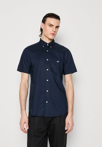 Man wearing a navy blue short-sleeve button-up shirt with a small logo on the chest, paired with black pants, standing against a plain background.