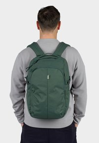 Zelený batoh s nastavitelnými popruhy, několika zipovými kapsami, hladkým textilním povrchem a nášivkou loga na přední straně.
