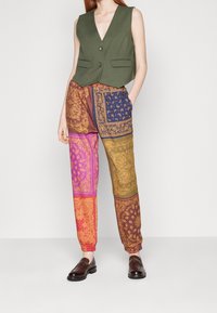 Chaleco sin mangas de color verde oliva con dos botones y bolsillos frontales, combinado con pantalones sueltos de múltiples patrones y colores que cuentan con puños elásticos.