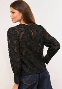 Crna bluza od čipke s cvjetnim uzorcima, prozirnim materijalom, dugim rukavima, detaljem na stražnjoj strani s prorezom i manžetnama na gumbe. Nosiva uz tamni traper.
