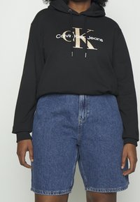 Svart huvtröja med "Calvin Klein Jeans"-logotyp i vitt och guld, tillsammans med blå denimshorts, som visar en avslappnad passform och modern stil.