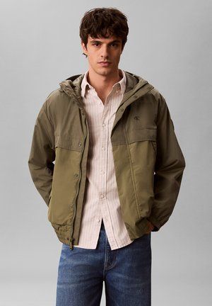 Jonge man met een olijfgroene capuchonjas over een licht gestreept overhemd met knopen en blauwe spijkerbroek, staand tegen een effen grijze achtergrond.