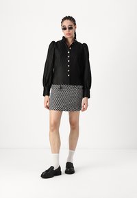 Svart plisserad blus med puffärmar och silverknappar, matchad med en randig minikjol. Tillbehör inkluderar svarta solglasögon och loafers.