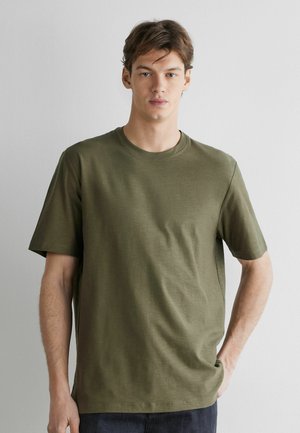 Jonge man met kort bruin haar, gekleed in een effen olijfgroen T-shirt met korte mouwen, staand tegen een lichtgrijze achtergrond.