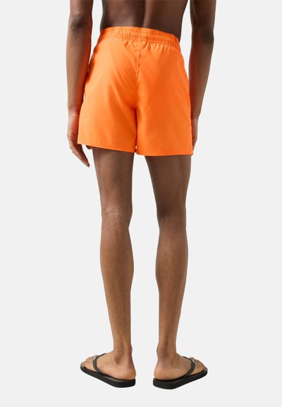 Man draagt felle oranje zwemshorts en zwarte teenslippers, staand met zijn rug naar voren tegen een effen achtergrond.