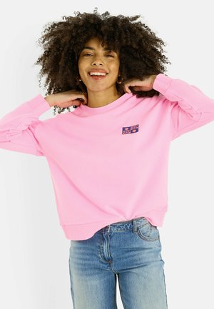 Femme souriante aux cheveux bouclés ajustant le col de son sweat-shirt rose orné de patches "LOVE", portant un jean bleu clair, sur fond blanc uni.