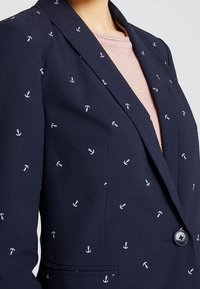 Námořnický blejzr s bílým vzorem kotvy, přiléhavý střih, klopené límce, zapínání na jeden knoflík a boční kapsy. Pod ním je texturovaný svetřík světle růžové barvy.