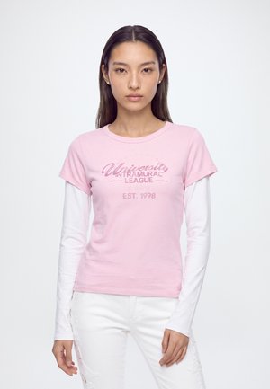 Giovane donna con lunghi capelli scuri indossa una maglietta rosa a maniche corte sopra una maglia bianca a maniche lunghe e pantaloni bianchi su uno sfondo uniforme.