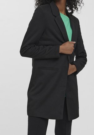 Femme portant un blazer noir par-dessus un haut vert, une main tenant le blazer fermé et l'autre dans la poche.
