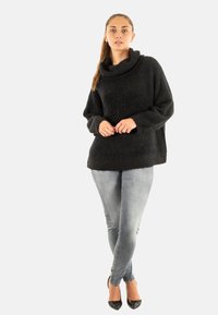 Frau steht in grauen Jeans, schwarzem Cowl-Neck-Pullover und schwarzen High Heels vor einfarbigem Hintergrund.