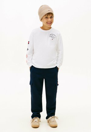 Chemise blanche à manches longues avec des graphiques imprimés, pantalon cargo bleu marine avec poches, bonnet en tricot beige, et chaussures de couleur claire.