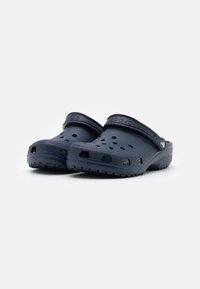Temno modri crocs z oblikovanim dizajnom, prezračevalnimi luknjami in teksturiranim podplatom za oprijem. Vključujejo hrbtni trak za varen oprijem.