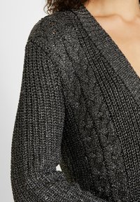 Cardigan en maille texturée noire avec un motif torsadé et des accents de fil métallique. Il présente un col en V profond et des manches larges.