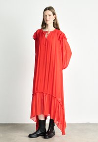 Vestido maxi rojo, fluido, con mangas con volantes y un dobladillo asimétrico, que presenta un detalle de lazo en el cuello. Llevado con botines negros.