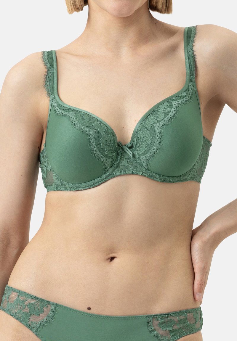 Reggiseno verde in pizzo con coppe sagomate, bordo in pizzo decorativo, spalline regolabili e un piccolo fiocco al centro. Design della schiena liscio.