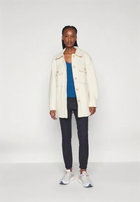 Bela ohlapna tkaninska jakna z velikimi gumbi, dvema sprednjima žepoma, kombinirana z modro majico in temnimi hlačami. Model nosi superge.