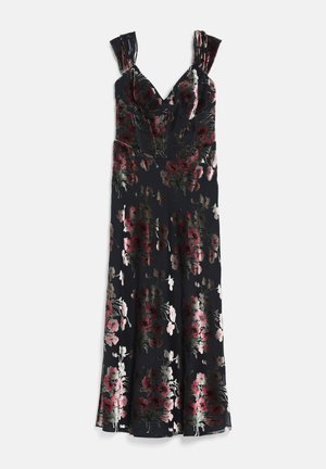 Donna che indossa un vestito nero a fiori con spalle scoperte e sottili bretelle, abbinato a tacchi neri e un braccialetto d'argento, in piedi su uno sfondo bianco.