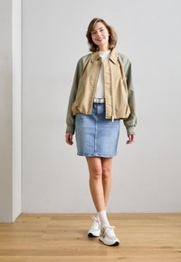 Giacca bomber color khaki e verde oliva, camicia bianca, gonna di jeans e sneakers con dettagli argento. Il modello è in piedi su un pavimento di legno, con uno sfondo neutro.